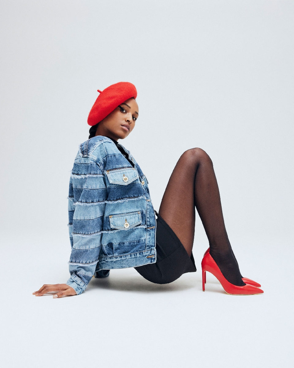 A fashionable woman in a denim jacket, red beret, and heels poses in a studio setting. The image highlights clothing and style.