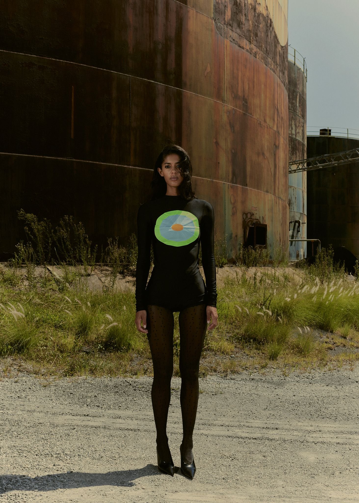 Adult female in long-sleeve dress posing in front of industrial tanks in outdoor setting with gravel ground and grassy vegetation.