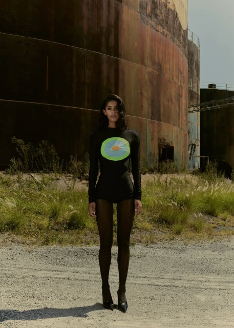 Adult female in long-sleeve dress posing in front of industrial tanks in outdoor setting with gravel ground and grassy vegetation.