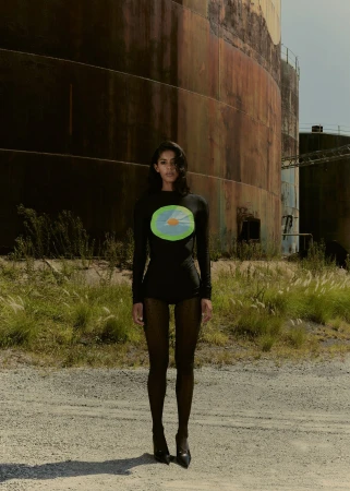 Adult female in long-sleeve dress posing in front of industrial tanks in outdoor setting with gravel ground and grassy vegetation.