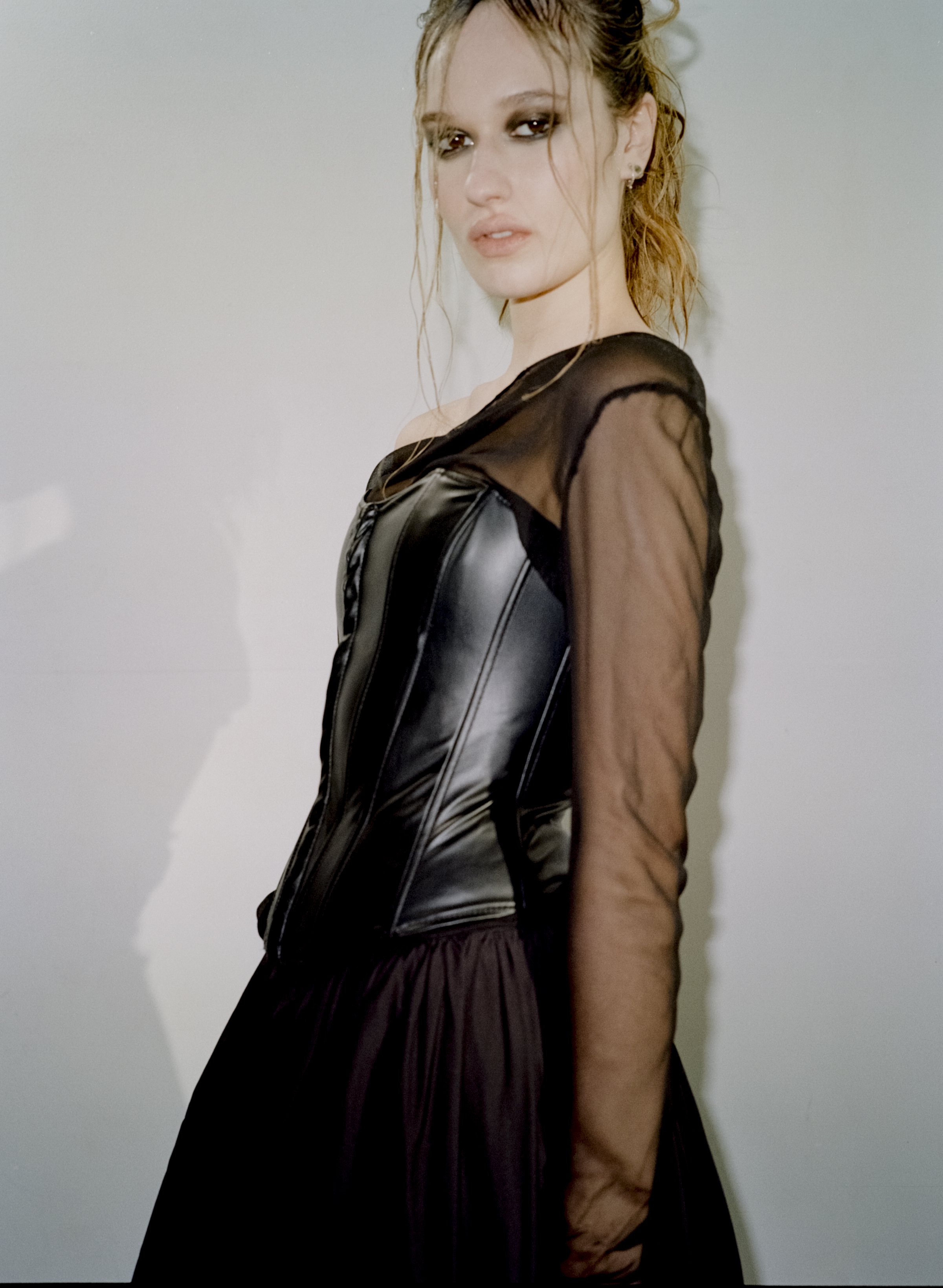 A stylish young adult woman with wet hair, wearing a black dress and corset, poses in front of a light grey studio backdrop.