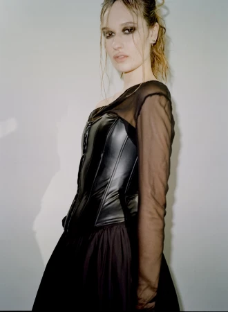 A stylish young adult woman with wet hair, wearing a black dress and corset, poses in front of a light grey studio backdrop.