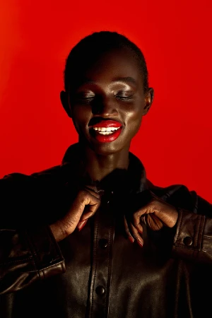 Close-up portrait of a smiling Black woman with bright red lipstick wearing a dark leather jacket against a bold red background.