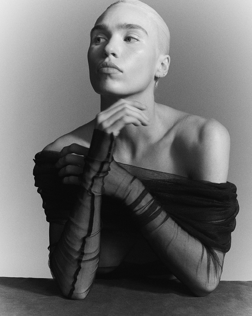 Black and white portrait of an androgynous person with shaved head, hands clasped, wearing sheer black fabric, gazing off-camera.