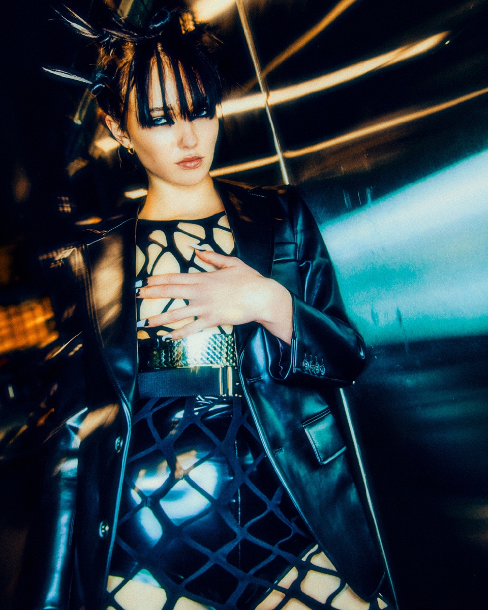Stylish woman in black coat and unique dress poses against blurred background, striking a confident pose with bold makeup.