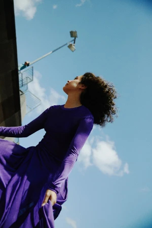 Low-angle shot of a woman with curly hair wearing a long purple dress against a bright blue sky with clouds.
