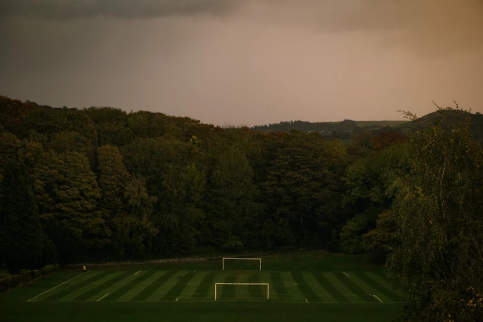 Overhead shot of a soccer field with neatly striped grass, surrounded by trees, under a cloudy sky, creating a serene sports landscape.