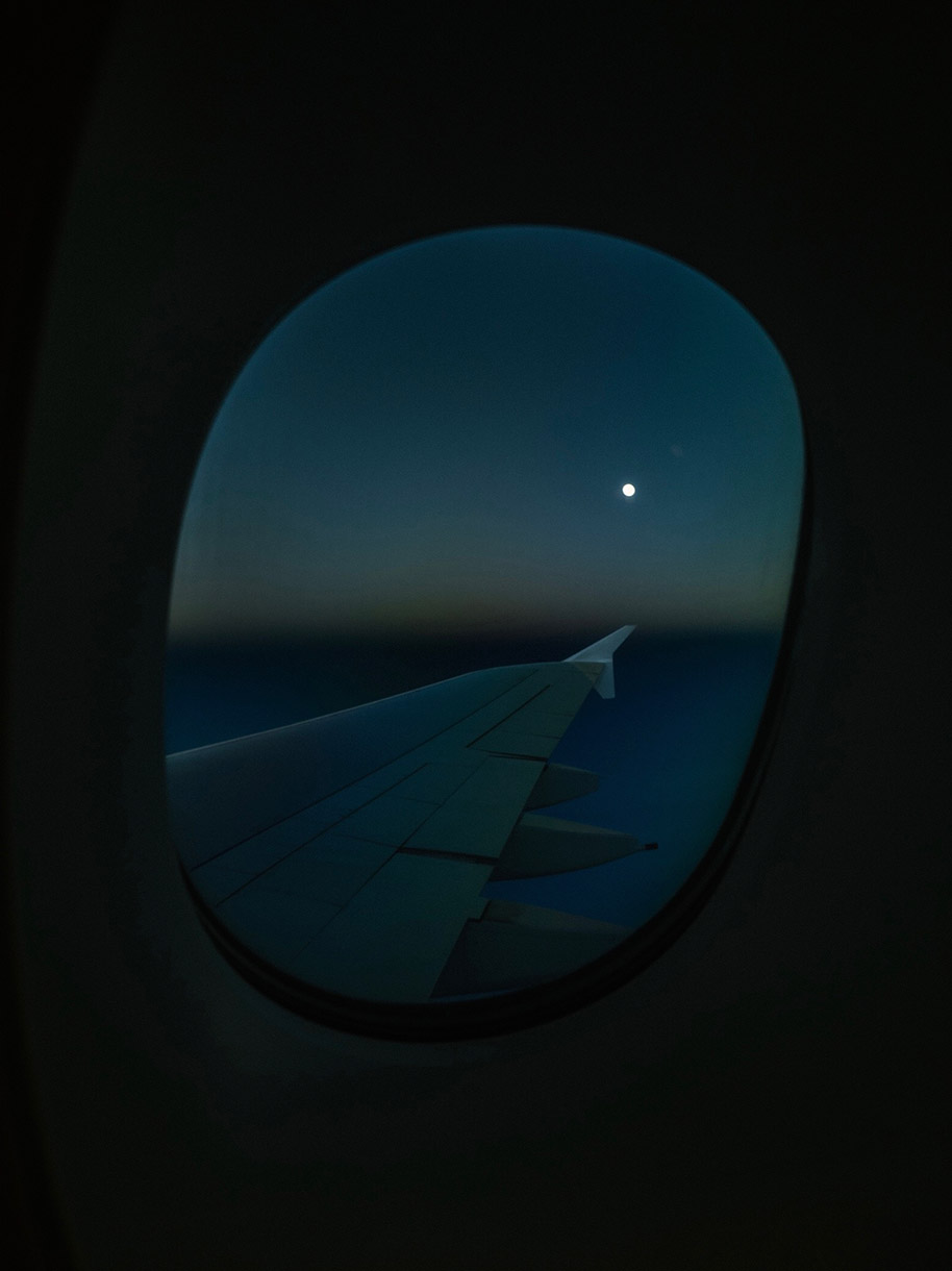 Airplane wing seen through a window at night, lit by the moon. The background is dark blue and black from the sky.