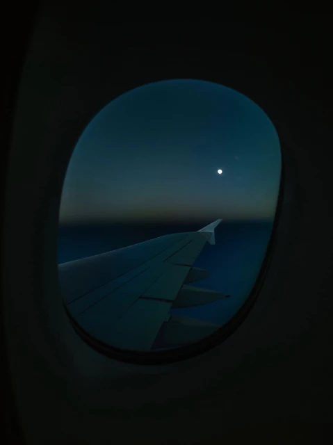 Airplane wing seen through a window at night, lit by the moon. The background is dark blue and black from the sky.