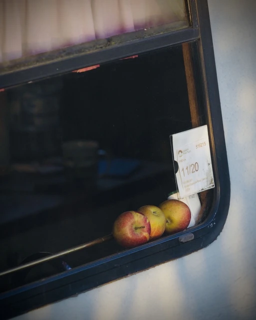 Three apples sit on the inside ledge of a camper van window, with light coming in from the window and a tag on the glass.