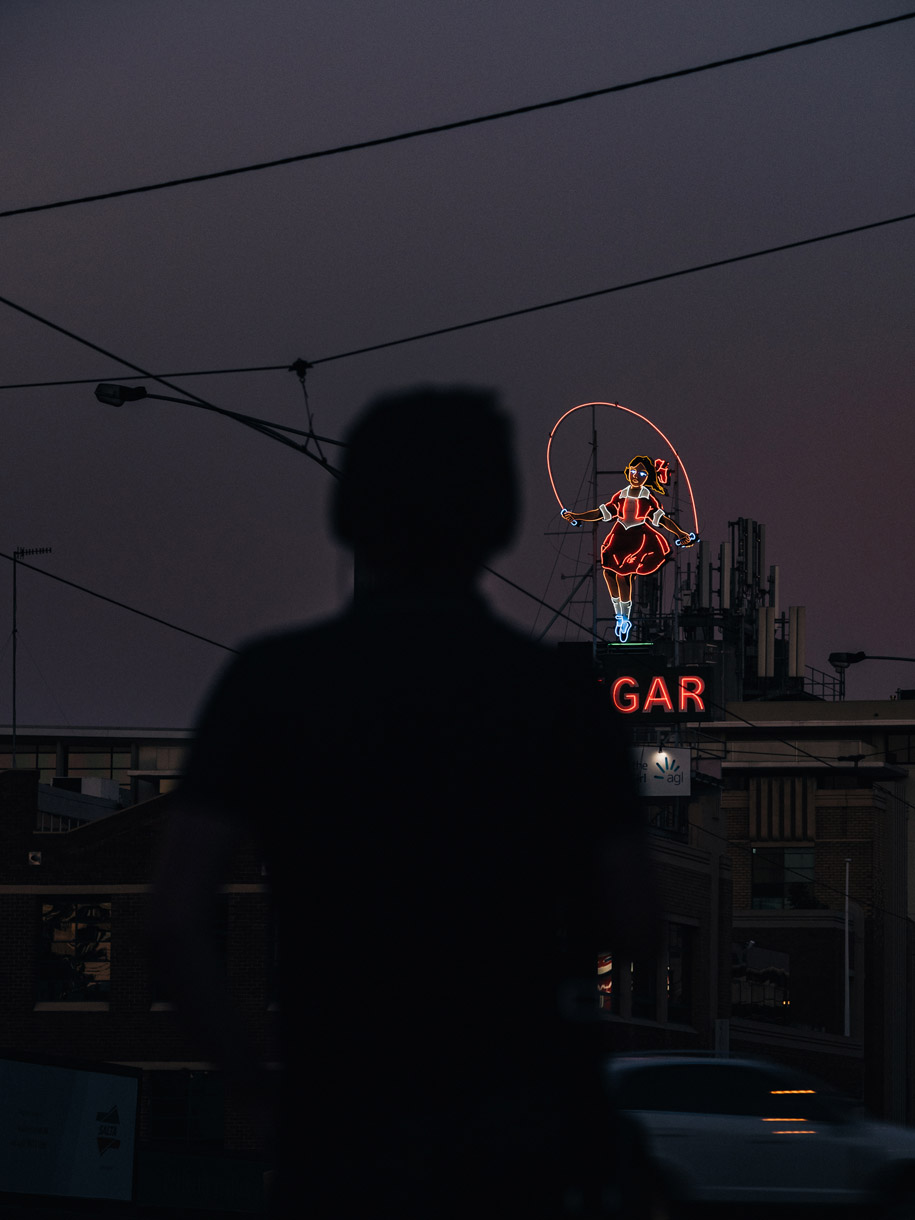 Person in shadow near neon sign of a girl jumping rope at night, dark and atmospheric, with building and wires in the background.