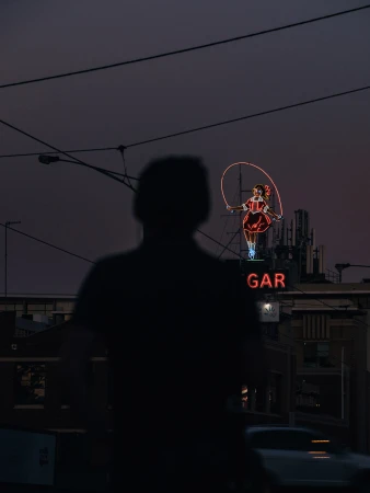 Person in shadow near neon sign of a girl jumping rope at night, dark and atmospheric, with building and wires in the background.