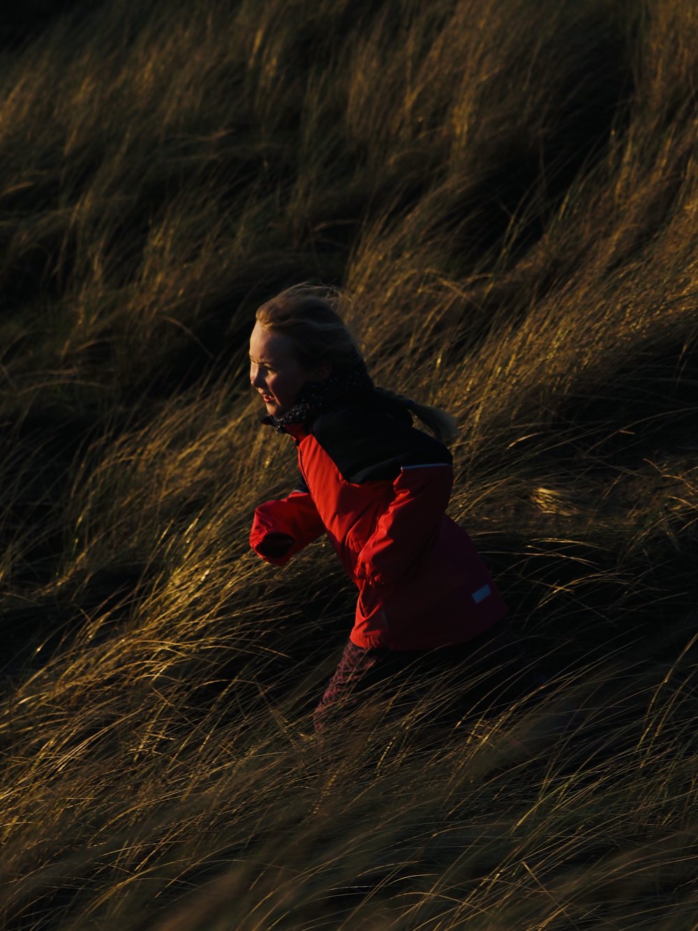 Child in red jacket running in tall grass in an outdoor setting, captured with a low angle view under natural lighting.