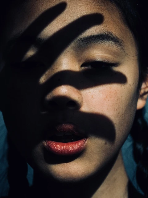 Close-up portrait of an Asian young adult female with hand shadow across her face, showing her dark hair, lips, and skin details.