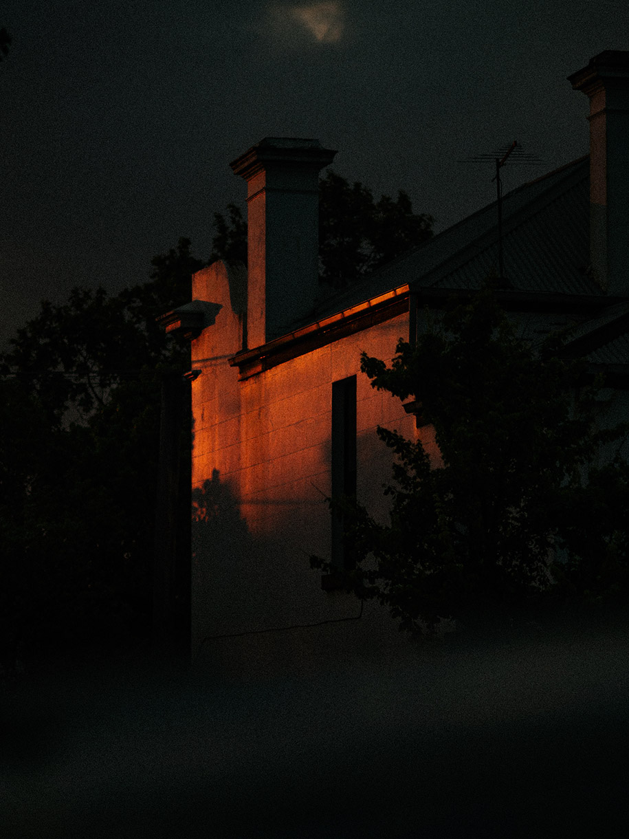 Orange light illuminates an old building with a chimney at night, framed by dark trees and sky. A serene, mysterious urban atmosphere.