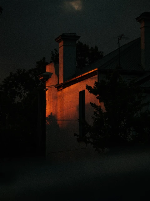 Orange light illuminates an old building with a chimney at night, framed by dark trees and sky. A serene, mysterious urban atmosphere.