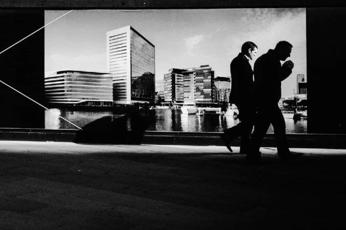 Two men in silhouette walking past a large urban waterfront mural in a high-contrast monochrome street photograph by Andy Hatton.