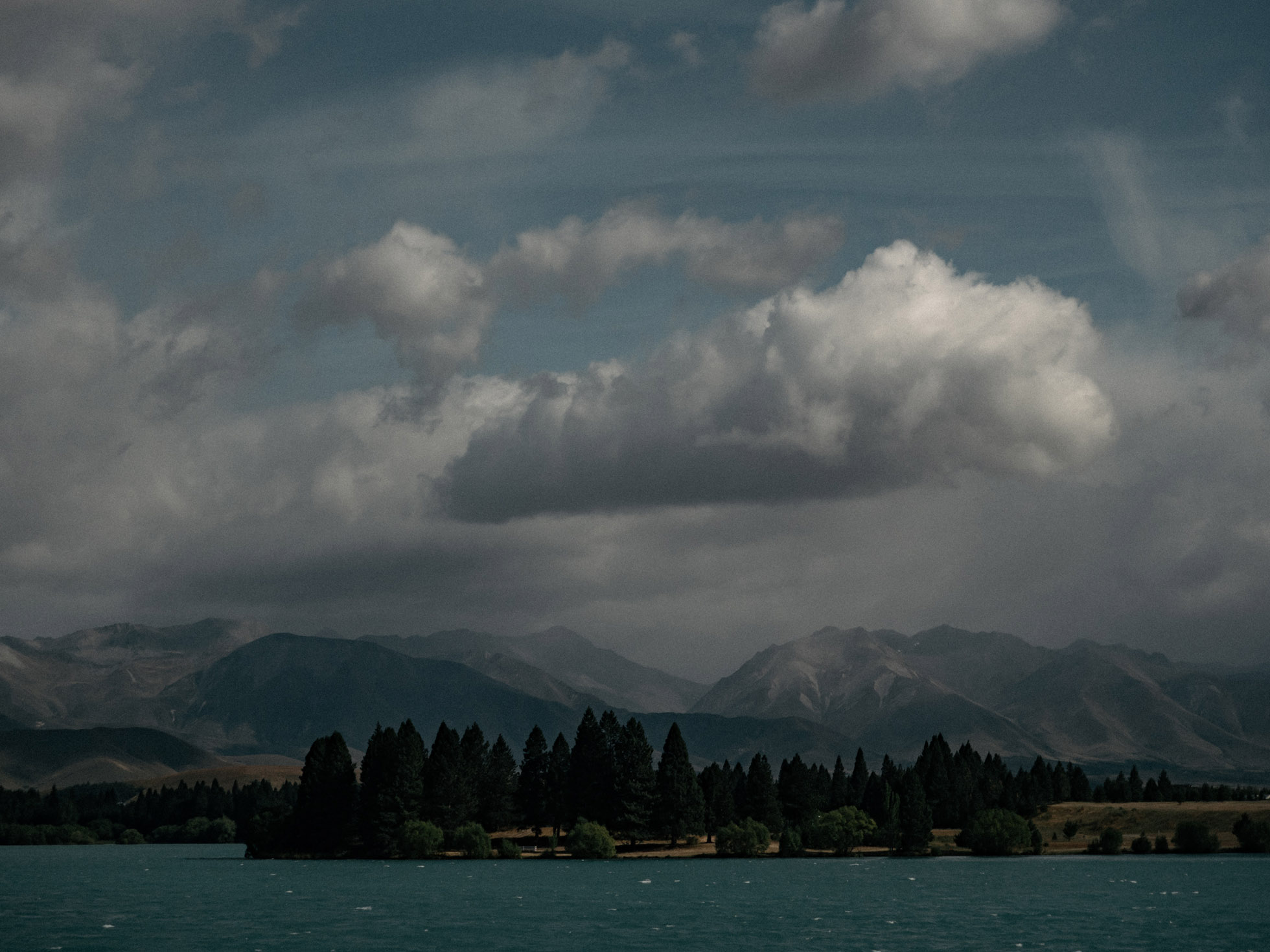 Lake Tekapo, New Zealand: a serene body of turquoise water reflecting a dark, forested shoreline under a cloudy sky, distant mountains.