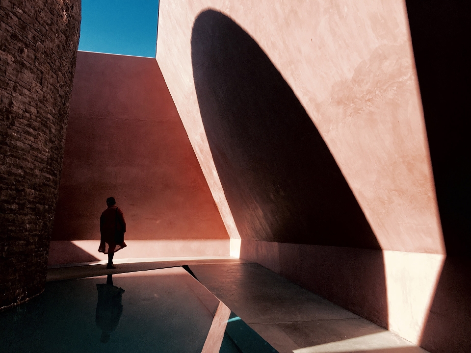 Person in red stands near the reflective water pool amidst the architectural elements of a pink building under a clear blue sky.