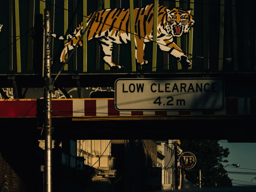Illustration of a tiger above a low clearance sign, with buildings visible in the background, captured during the daytime.