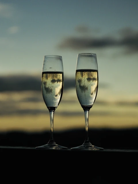Two champagne flutes filled with sparkling wine stand on a dark surface, with a blurred sky and landscape in the background.