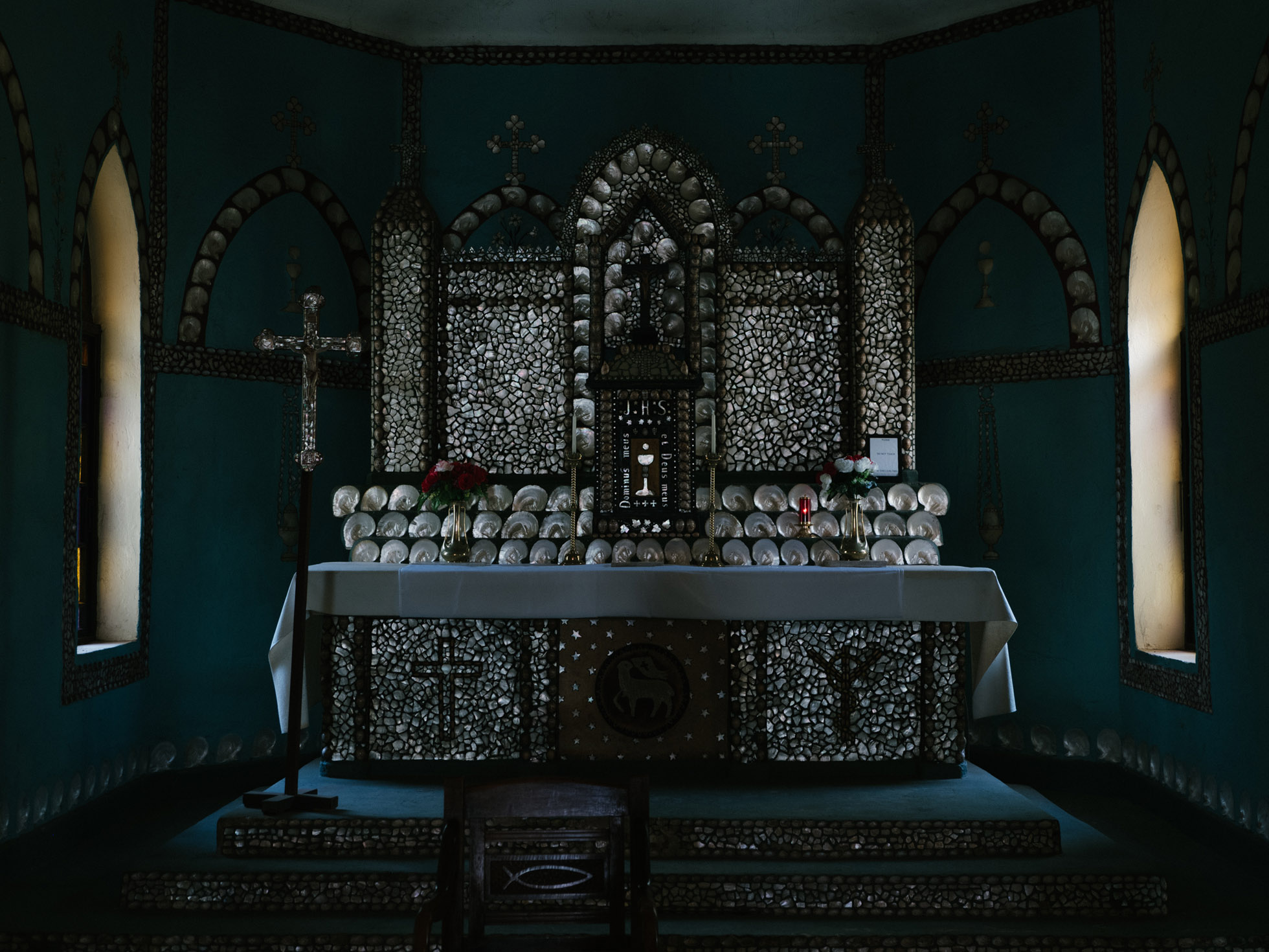 Interior view of a chapel featuring an altar, crucifix, and decor with numerous seashells and floral arrangements, illuminated by natural light.