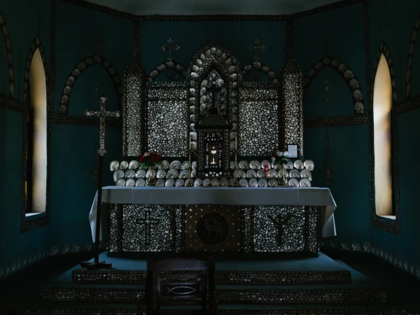 Interior view of a chapel featuring an altar, crucifix, and decor with numerous seashells and floral arrangements, illuminated by natural light.