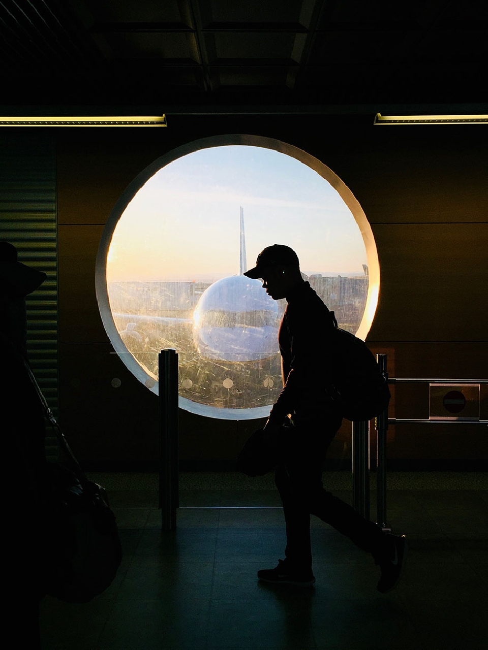 Silhouette of a person walking inside an airport terminal with a large round window showing an airplane at sunset.