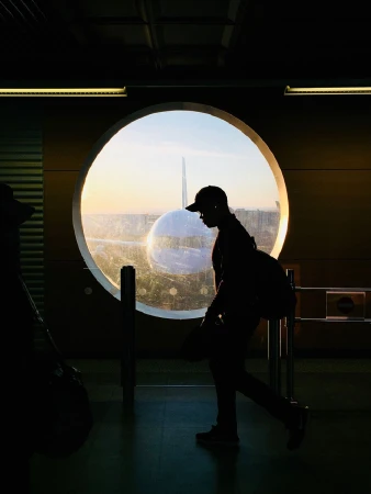 Silhouette of a person walking inside an airport terminal with a large round window showing an airplane at sunset.