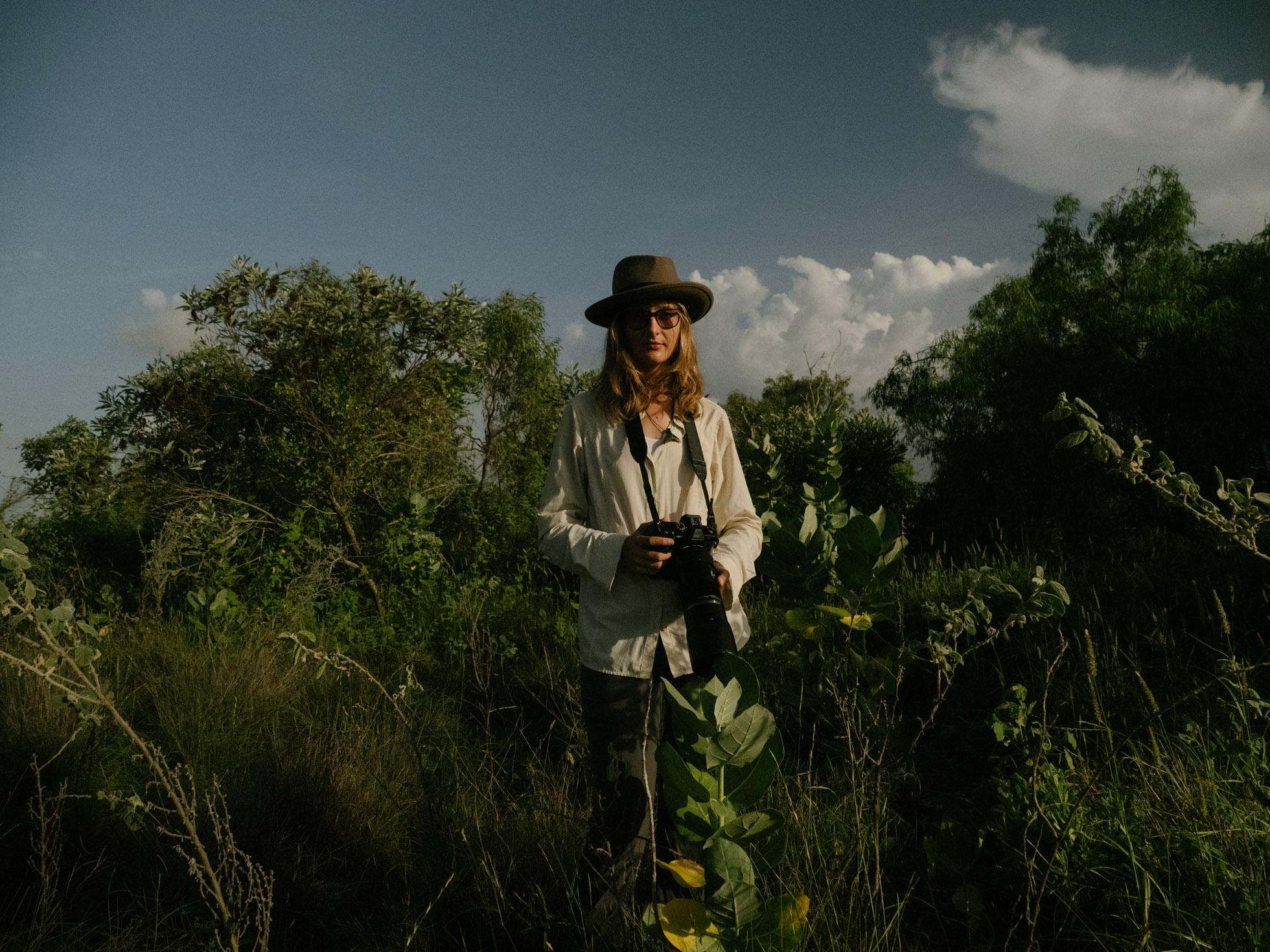 Adult female nature photographer stands in a lush field with a camera, wearing a hat and sunglasses, under a partly cloudy sky.