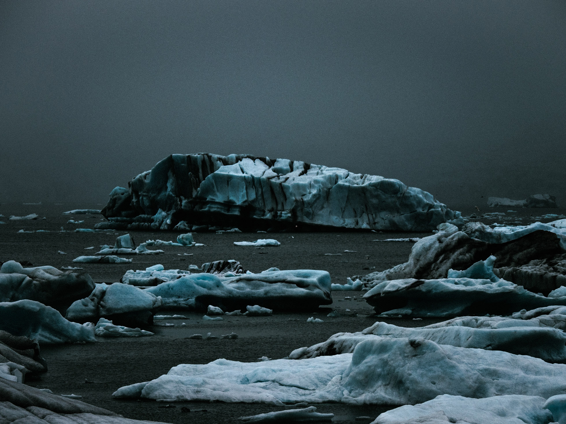 Icebergs dot a dark, frigid body of water under a cloudy sky, creating a stark, remote Arctic landscape with dramatic lighting.