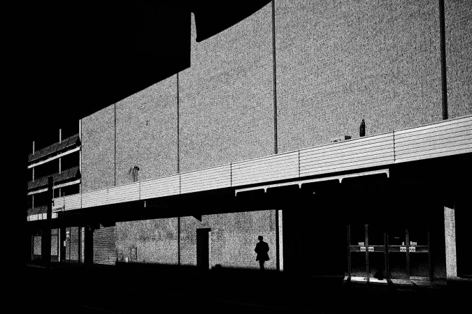 High-contrast monochrome photograph of a lone figure walking beneath the facade of a large commercial building at night.