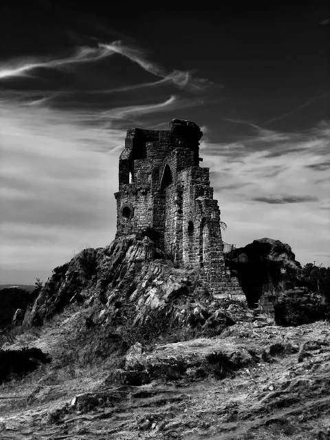 Black and white photo of castle ruins atop a grassy hill against a cloudy sky, showcasing architectural detail and natural landscape.