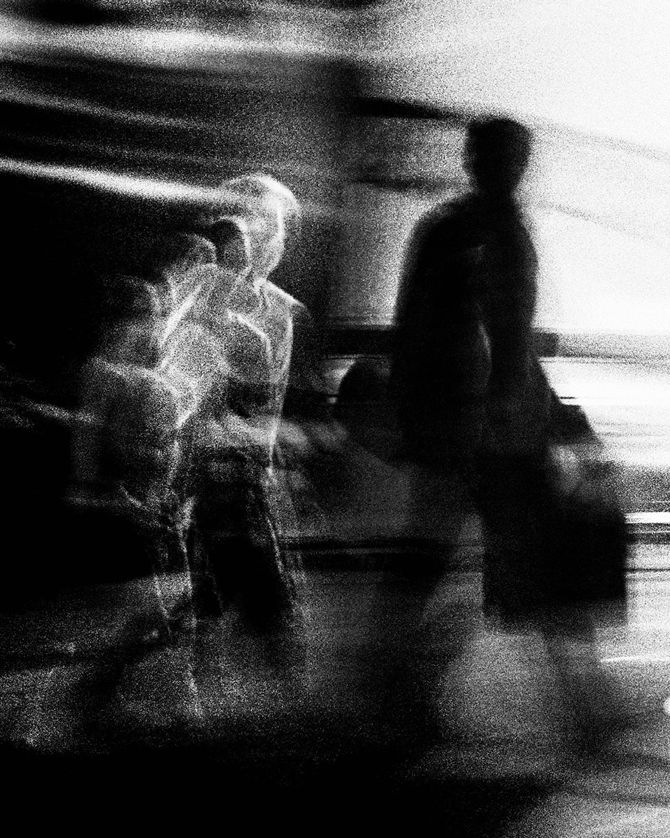 Monochrome motion blur of two adult males walking in an indoor setting. One is in the foreground, and one is behind him.