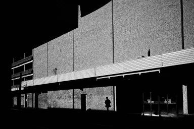 High-contrast monochrome photograph of a lone figure walking beneath the facade of a large commercial building at night.