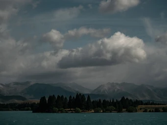 Panoramic landscape photograph of a dark turquoise lake with a line of dark pine trees beneath distant mountains under a cloudy sky.