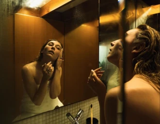 Woman with wet hair wrapped in a towel looking into a bathroom mirror, photographed by Andy Hatton.