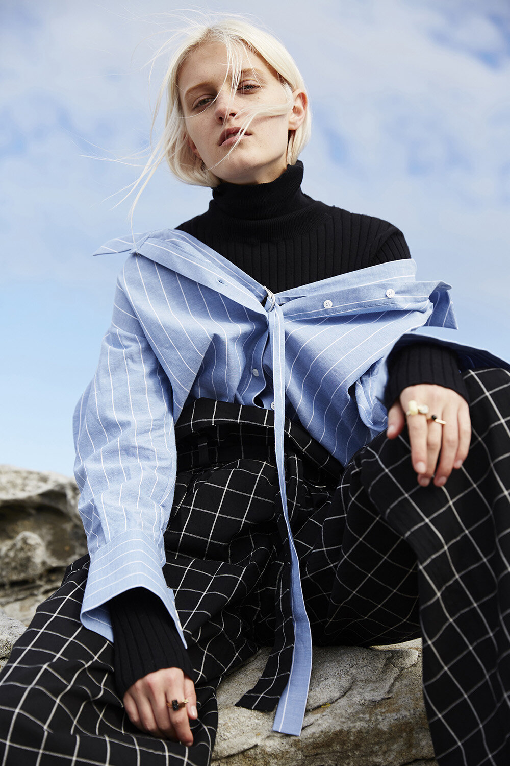A fair-skinned woman in a black top and blue shirt poses on a rocky surface against a light blue sky, showcasing fashion apparel.