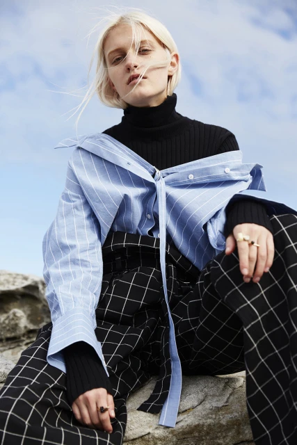 A fair-skinned woman in a black top and blue shirt poses on a rocky surface against a light blue sky, showcasing fashion apparel.