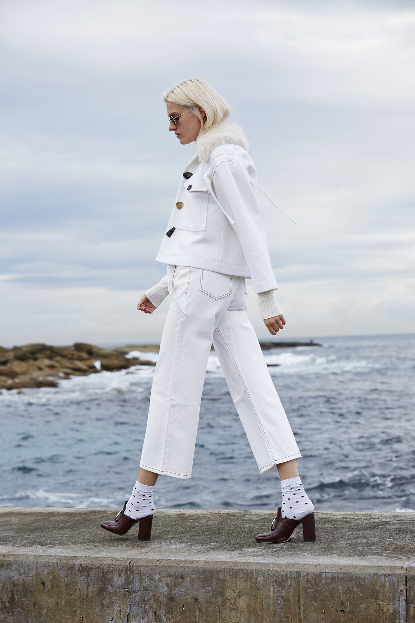Fashionable woman in a white denim jacket and jeans walks along a concrete ledge by the ocean on an overcast day.