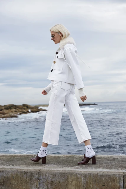 Fashionable woman in a white denim jacket and jeans walks along a concrete ledge by the ocean on an overcast day.