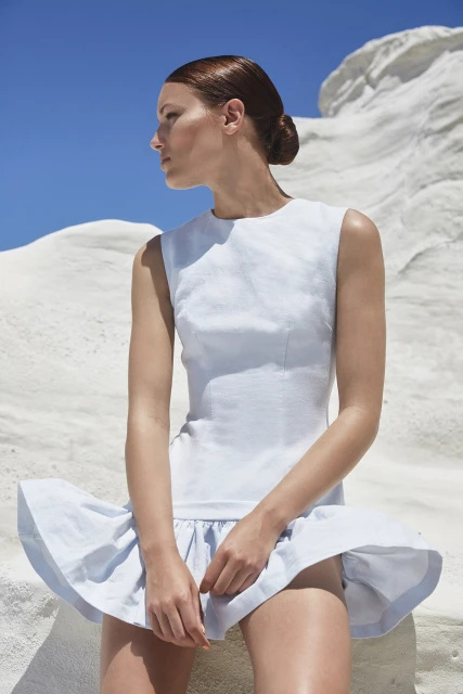 A woman in a light blue dress poses in front of a white rock formation with a clear blue sky in an outdoor fashion shot.