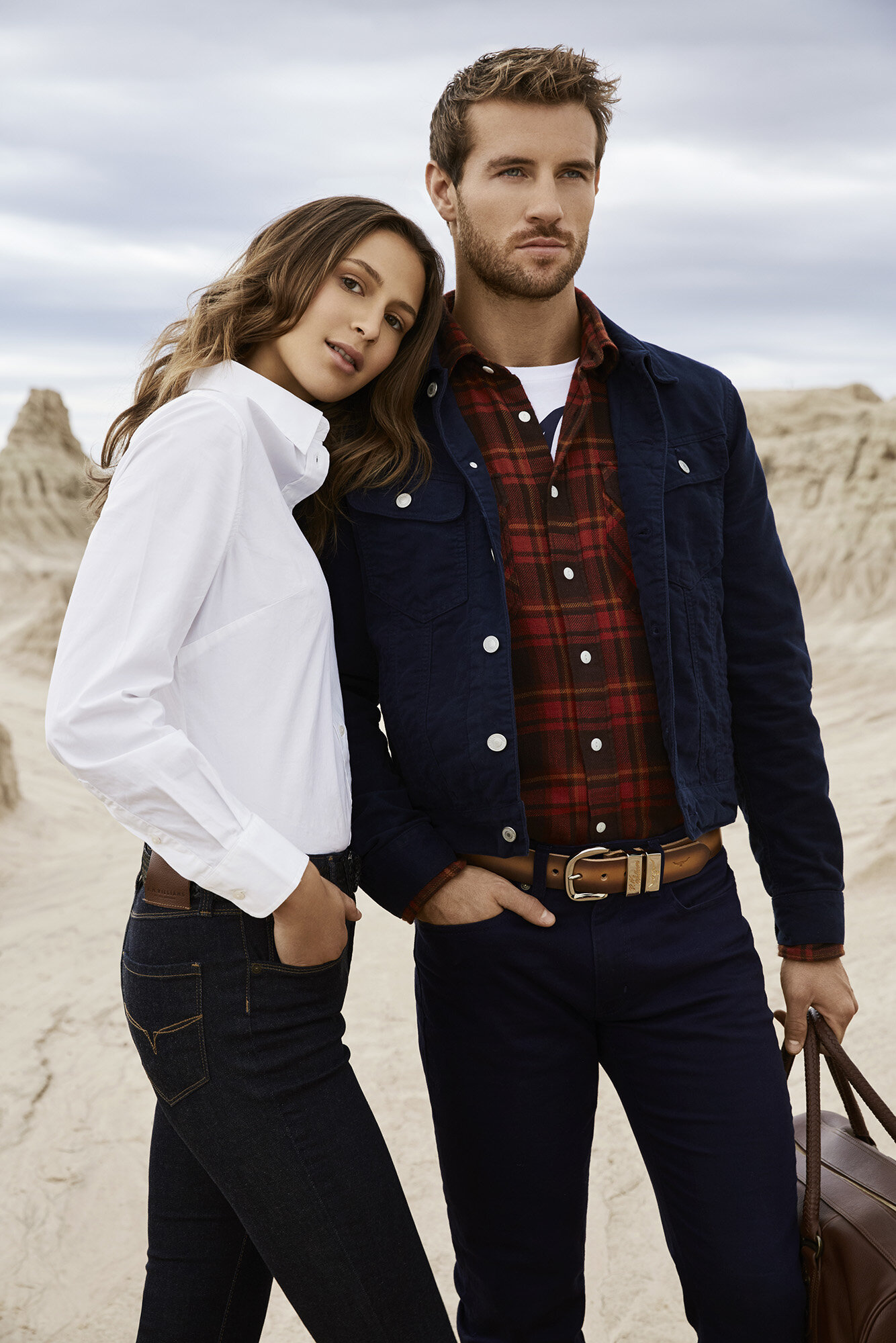 A stylish couple poses outdoors in a promotional fashion shot. The adult male and female wear complementary casual attire and leather belts.