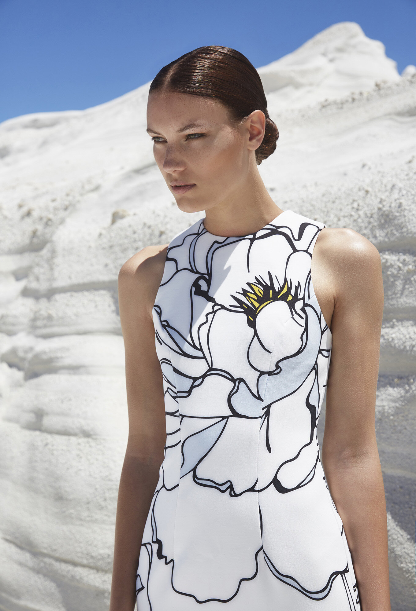 Woman in white floral dress posing outdoors against a textured white background under a bright blue sky.