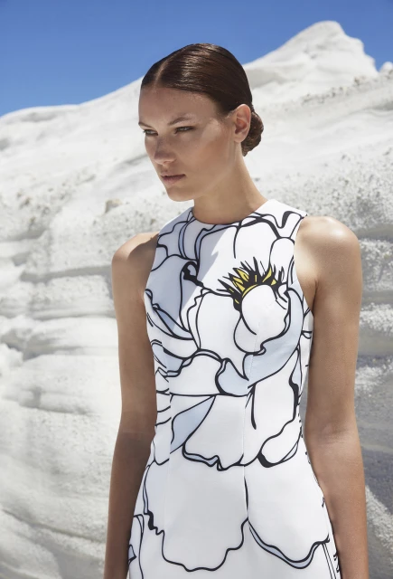 Woman in white floral dress posing outdoors against a textured white background under a bright blue sky.