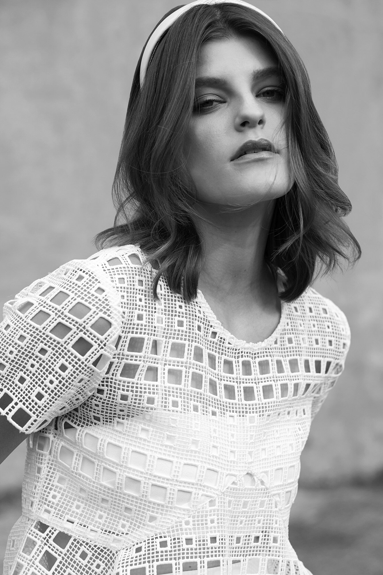 Monochrome portrait of a woman with shoulder-length hair, wearing a headband and white top with patterned cutouts against a plain backdrop.