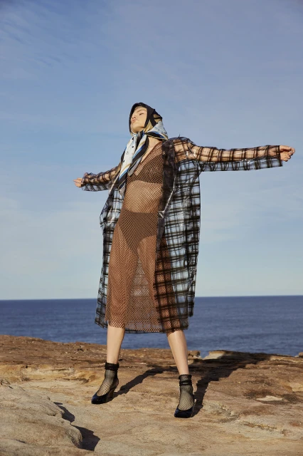 Fashionable woman in sheer plaid coat on rocky coast with ocean and sky. Sheer dress, headscarf, and ankle boots complete the look.