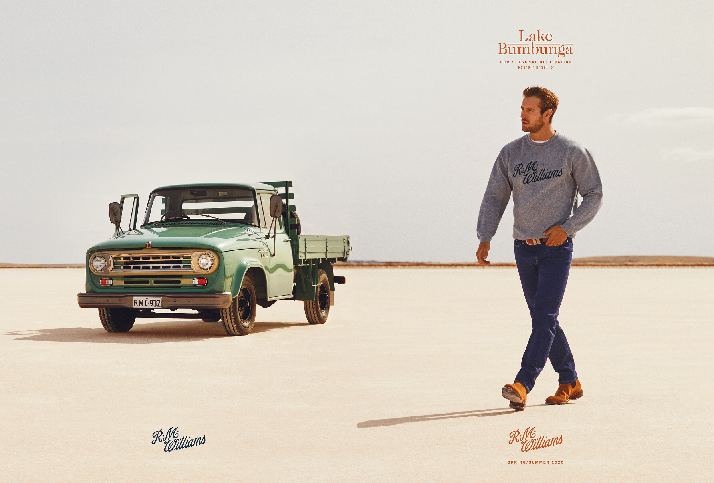 An adult male walks across a desert landscape, wearing a gray sweater and jeans. A green vintage truck is in the background.