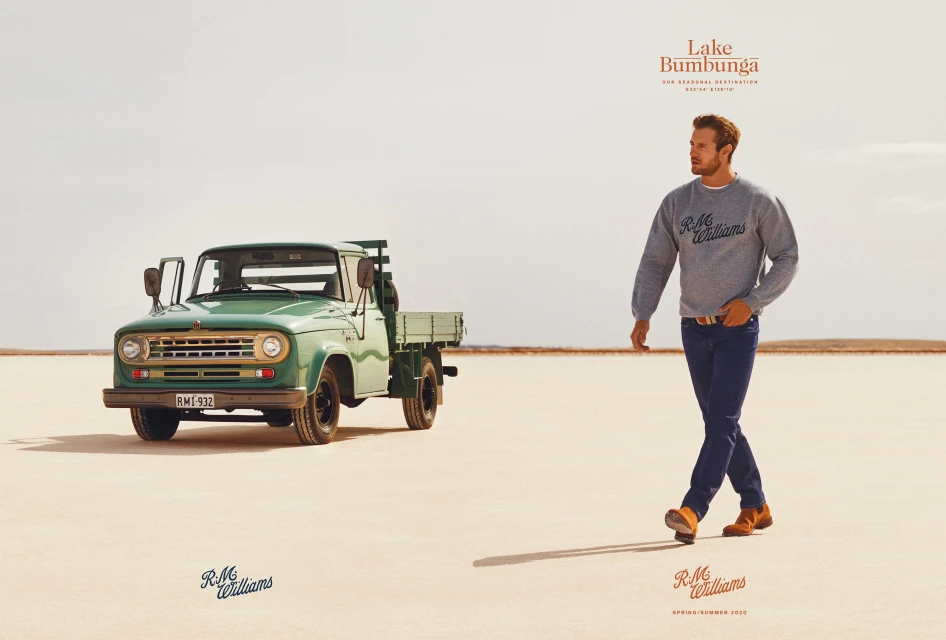 An adult male walks across a desert landscape, wearing a gray sweater and jeans. A green vintage truck is in the background.