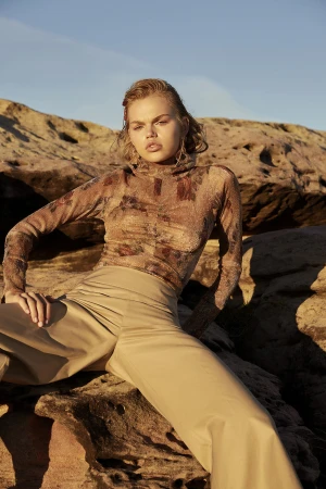 Model in a floral sheer turtleneck and beige trousers seated against rocky sandstone cliffs under warm sunlight.